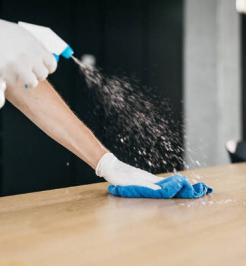 A close-up shot of cleaning a wooden desk in an office with a disinfectant to limit the spread of COVID-19. Only hands wearing medical gloves are visible, no faces. Horizontal daylight indoor photo.
